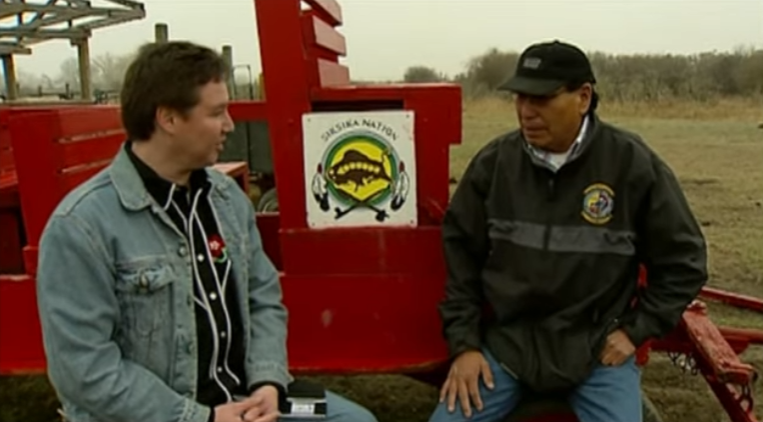 Screenshot 2025-12-02 184457 Two men having a conversation beside some heavy machinery in a field.