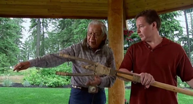 Screenshot 2025-12-02 190945 An elder teaching a man how to use a wooden tool while in a gazebo like structure.