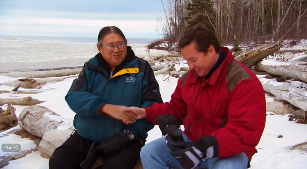 Screenshot 2025-12-02 191612 Two men sitting on a log near a body of water in the winter having a conversation.