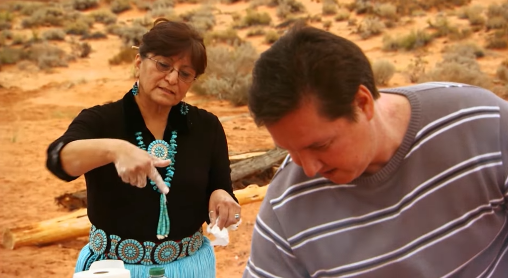 Screenshot 2025-12-02 191913 A woman intrusting a man on how he is cooking food over a campfire.